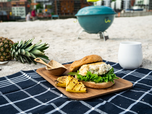 Strand med ananas i bakgrunnen, en hvit plastkopp, et blått håndkle med en fiskeburger oppå og grillet ananas. 