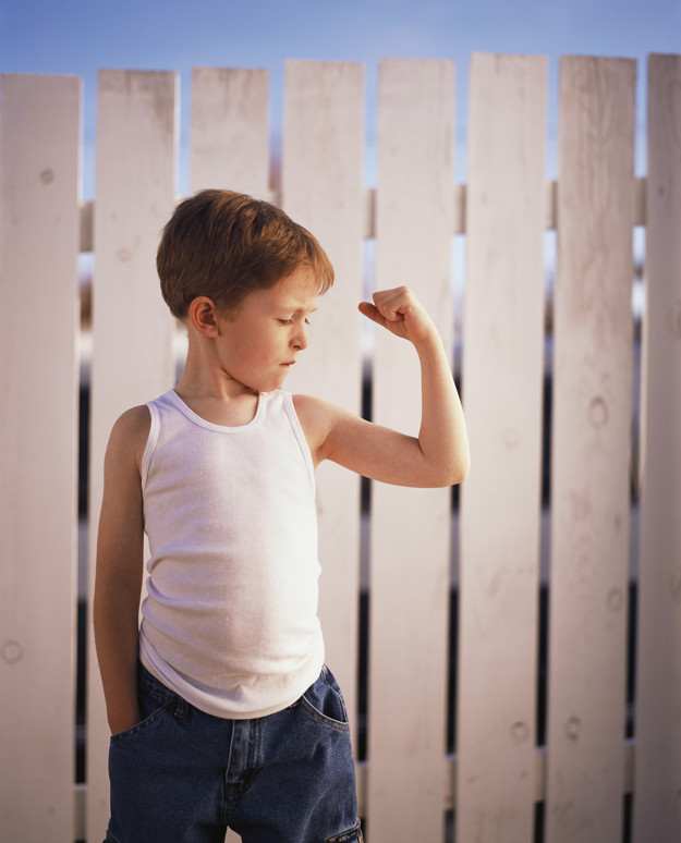 En ung gutt som flekser ene armen. Han går i mørk jeans og hvit singlet. Bak han er hvitt stakittgjerde. 